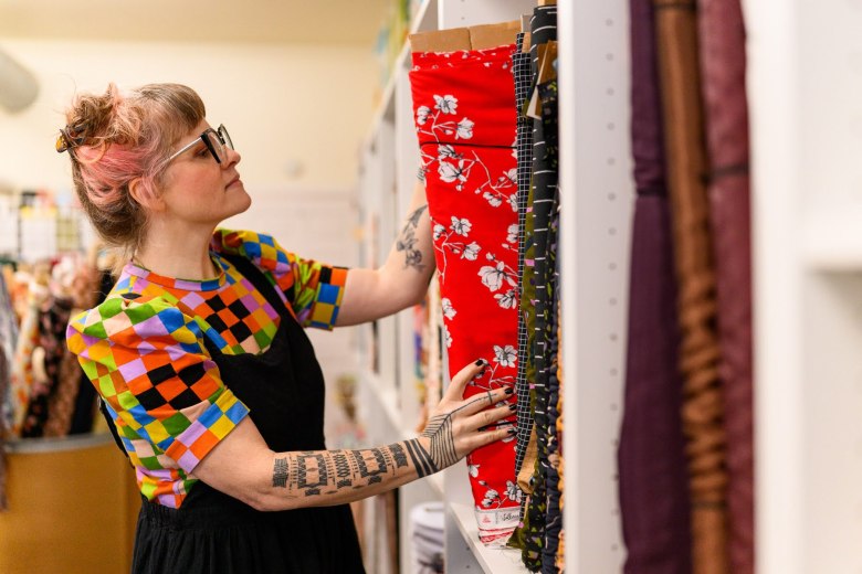 A person with glasses and tattoos selects a bolt of red floral fabric from a shelf in a fabric store.