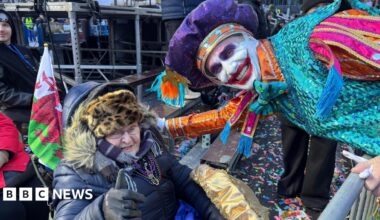 Photograph of Avril Davidge, sat outside in her wheelchair. A Welsh flag can be seen poking out of her wheelchair. She wears a leopard furry hat, a purple scarf and a black puffer coat. She wears small reading glasses and holds her thumb up to the camera. A captain of the Mummers parade leans down to her and smiles. He wears a vibrant blue, purple and orange sequined costume and has a large red joker smile painted on his face.