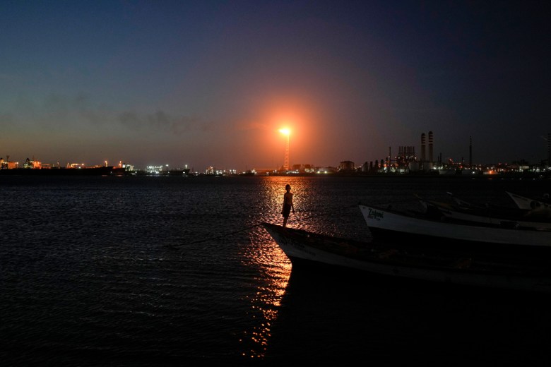 A person stands on the bow of a boat at dusk, with an industrial facility and a bright flare in the background across the water.