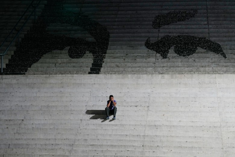 A person sits alone on wide concrete steps beneath a large artistic depiction of two eyes on the upper stairs.