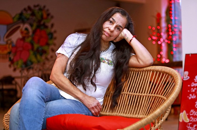 A woman with long dark hair sits on a wicker chair with a red cushion, resting her head on her hand, in a room decorated with red lights and colorful wall art.