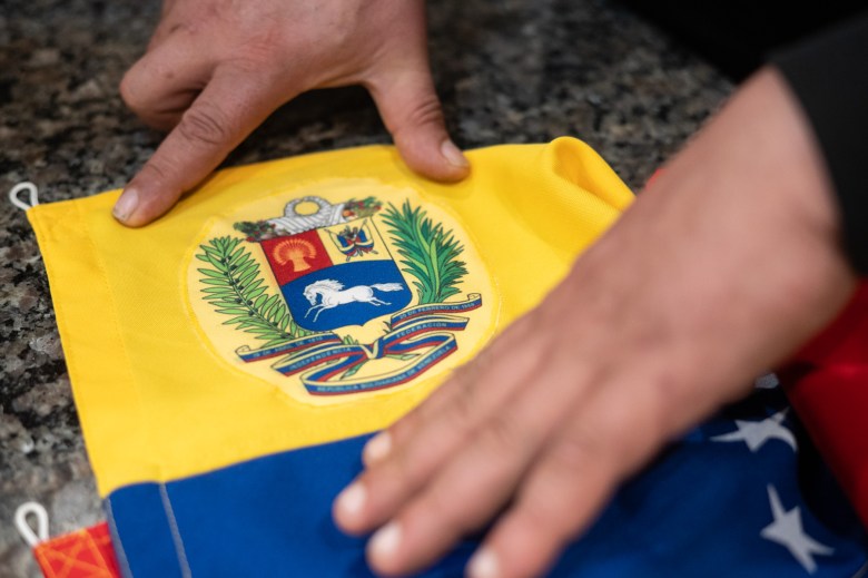 Two hands touch a Venezuelan flag laid on a speckled countertop, focusing on the national coat of arms in the yellow section.