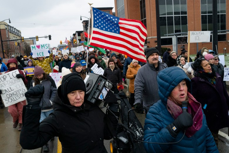 A group of people march on a city street holding protest signs and a large American flag; one person uses a megaphone and another carries a video camera.