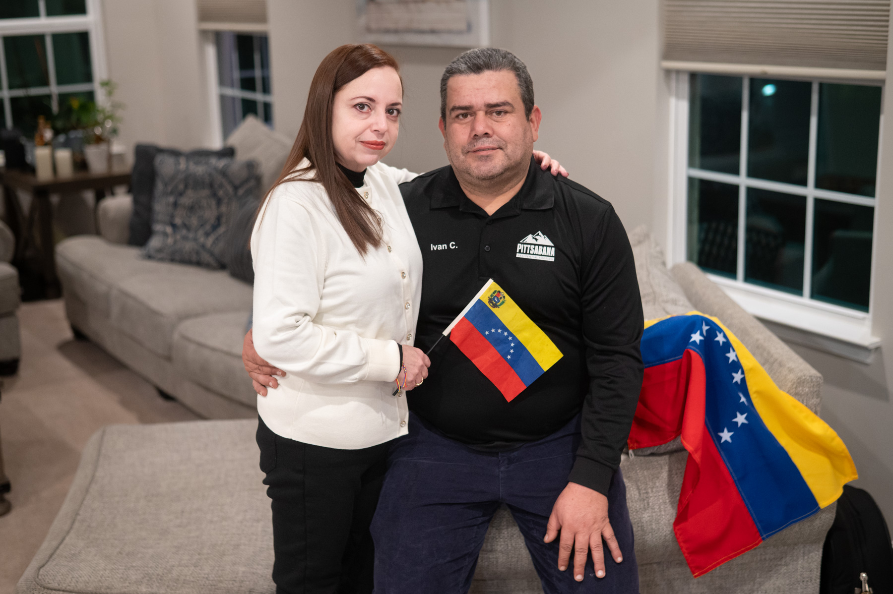 A woman and a man sit together on a sofa; the man holds a small Venezuelan flag, and a larger Venezuelan flag is draped on the couch beside them.