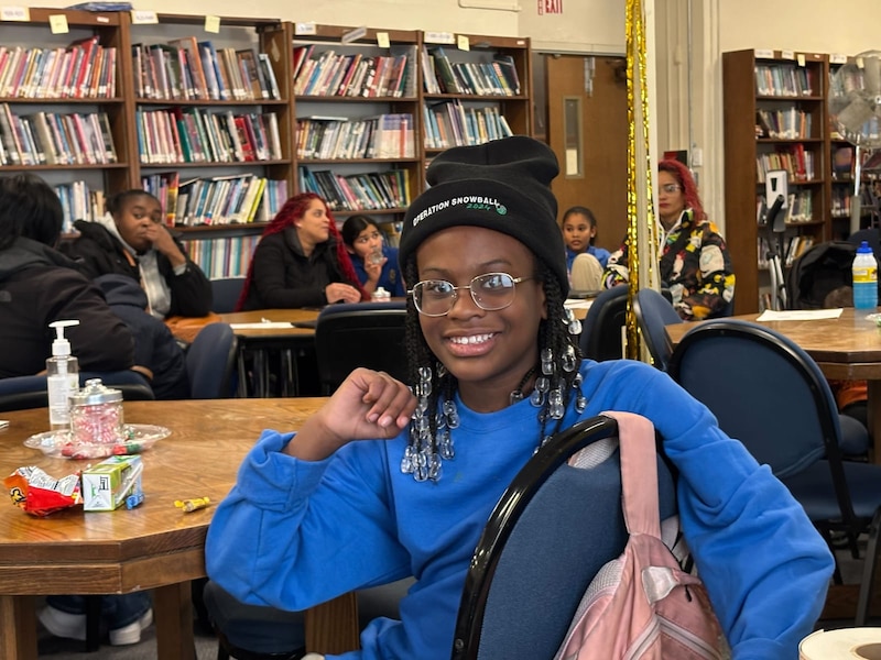 A Black teenage girl in a blue sweater and wearing a black hat poses for a portrait while smiling and sitting at a wooden desk in a classroom.
