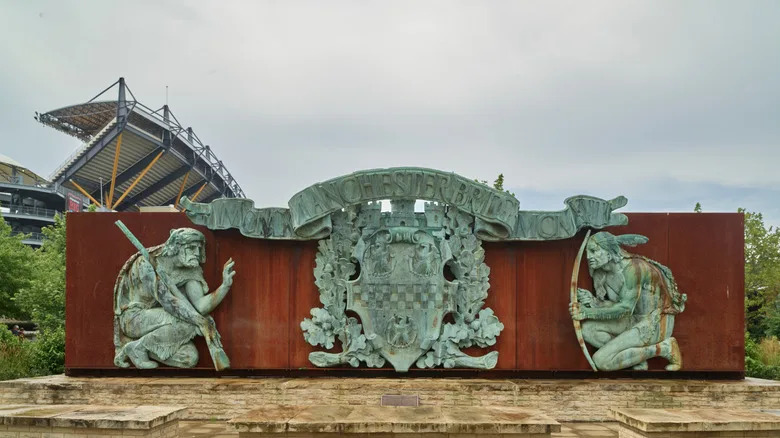 Manchester Bridge statue in Pittsburgh's North Side, with bronze figures against a red surface with Acrisure Stadium in the background, under a cloudy sky