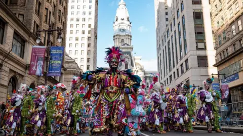 Visit Philadelphia Photograph showing a crowd of people in elaborate costumes are picturing walking down Broad Street in Philadelphia. In the centre, the captain can be seen in a multicoloured costume, with large pink feathers poking out of a head piece. 