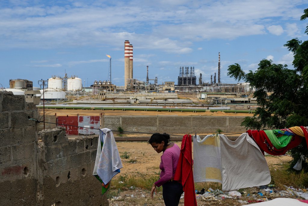 A woman hangs laundry on a clothesline near a crumbling wall, with an industrial refinery and smokestacks in the background under a partly cloudy sky.