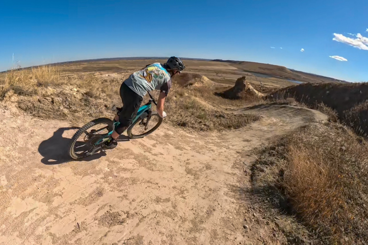 A person riding a mountain bike along a dirt trail in a vast, open landscape with gentle hills and blue sky. The cyclist is leaning slightly to navigate a curve on the path, with dry grass and sparse vegetation visible on the sides.