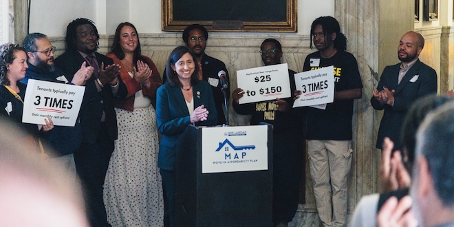 Surrounded by supporters holding signs bearing information about rental housing costs, Councilmember At-Large Rue Landau, a petite White woman with dark brown hair in a blue suit, applauds and smiles.