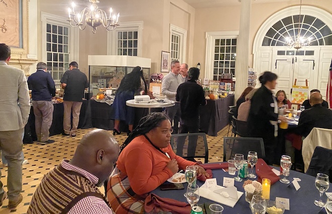 A group of people sit in a dining room in a historic feel, dining, talking, and serving themselves from a buffet.