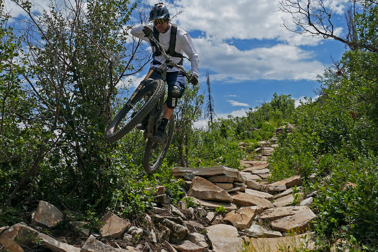 A mountain biker jumps over a rocky trail surrounded by greenery under a blue sky with scattered clouds. The rider is wearing a helmet and protective gear, showcasing an action-packed moment in outdoor sports.