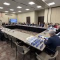 About two dozen people sit at tables set in the form of a rectangle in a conference room.