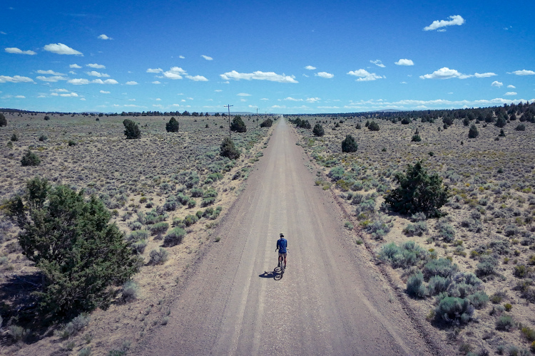 A cyclist riding on a dirt road through a wide, open landscape with sparse vegetation and a clear blue sky scattered with clouds. Power lines run alongside the road, leading into the distance.