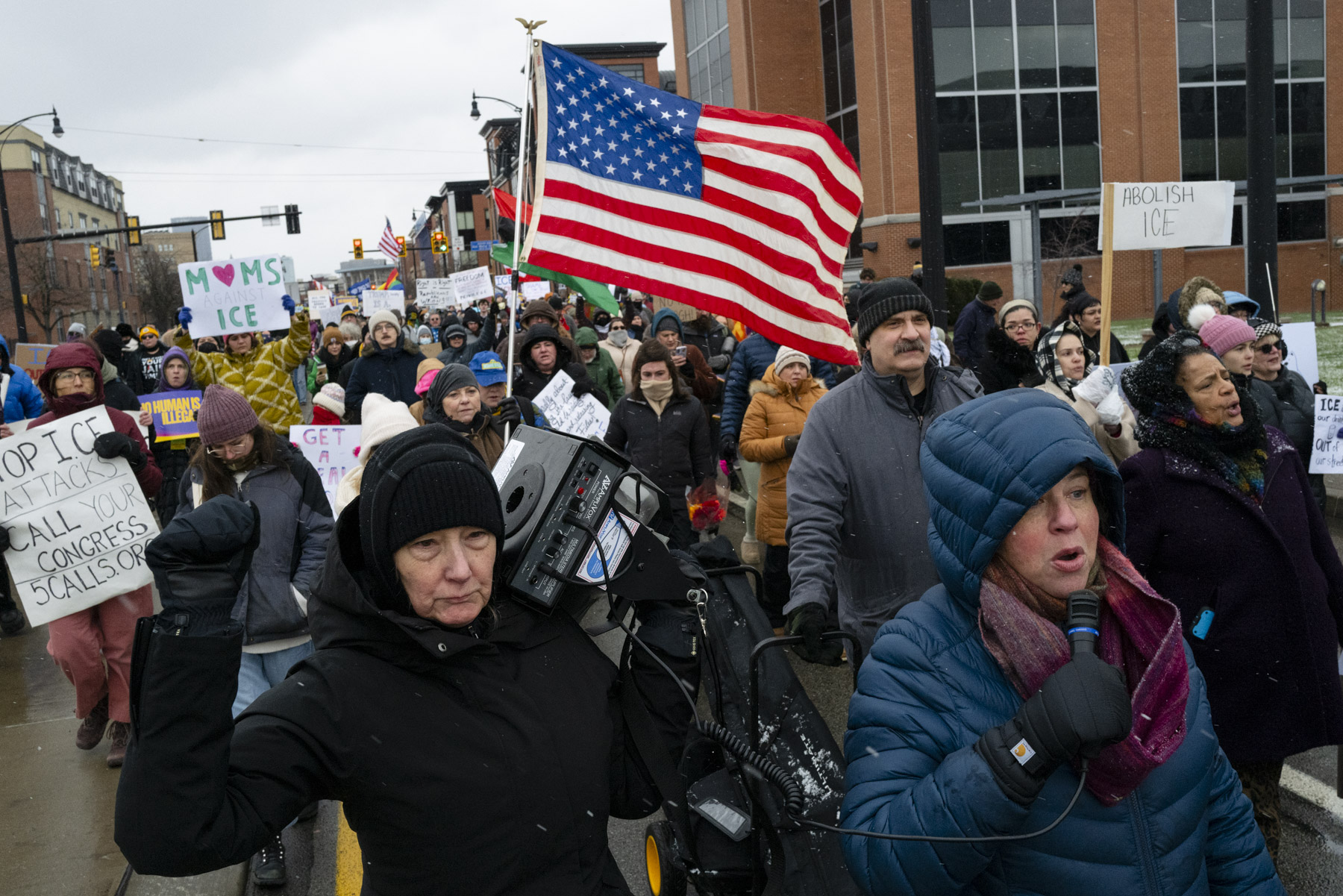 A group of people march on a city street holding protest signs and a large American flag; one person uses a megaphone and another carries a video camera.