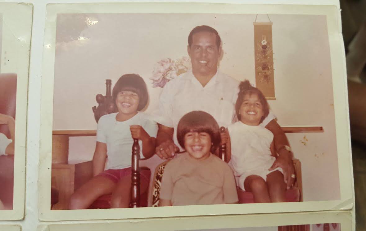 A man sits on a chair with three smiling children, two girls and one boy, posing indoors in front of a wall with a hanging decoration and a flower arrangement.