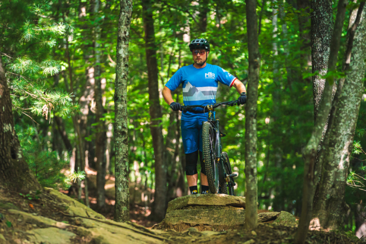 A mountain biker in blue athletic gear stands on a rock in a wooded area, holding his bike. The surrounding forest features tall trees and lush greenery, creating a vibrant outdoor setting.