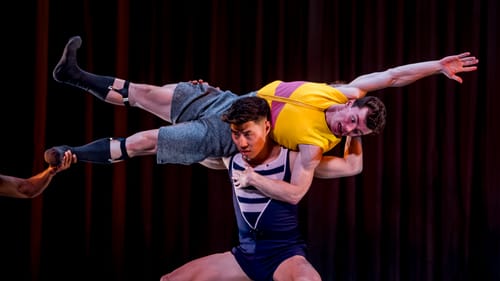 Closeup on two muscular male dancers, one holding the other’s straightened body across his shoulders, parallel to the floor