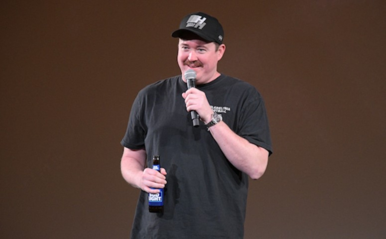 shane gillis talking on stage in a black shirt holding a beer