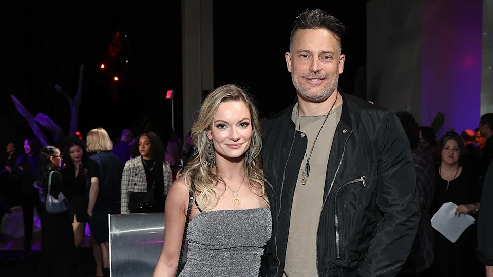 Caitlin O'Connor wearing a sleeveless grey dress standing next to Joe Manganiello wearing a black jacket and a grey shirt.