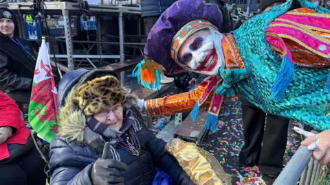 Kory Aversa Photograph of Avril Davidge, sat outside in her wheelchair. A Welsh flag can be seen poking out of her wheelchair. She wears a leopard furry hat, a purple scarf and a black puffer coat. She wears small reading glasses and holds her thumb up to the camera. A captain of the Mummers parade leans down to her and smiles. He wears a vibrant blue, purple and orange sequined costume and has a large red joker smile painted on his face. 