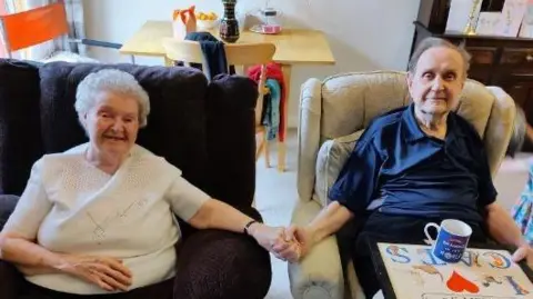 Fiona Davidge Photograph of Avril (left) and Clifford (right) sat on two armchairs. They hold each other's hands in the centre of the image and smile at the camera. Avril wears a white t-shirt, while Clifford wears a navy polo shirt. 