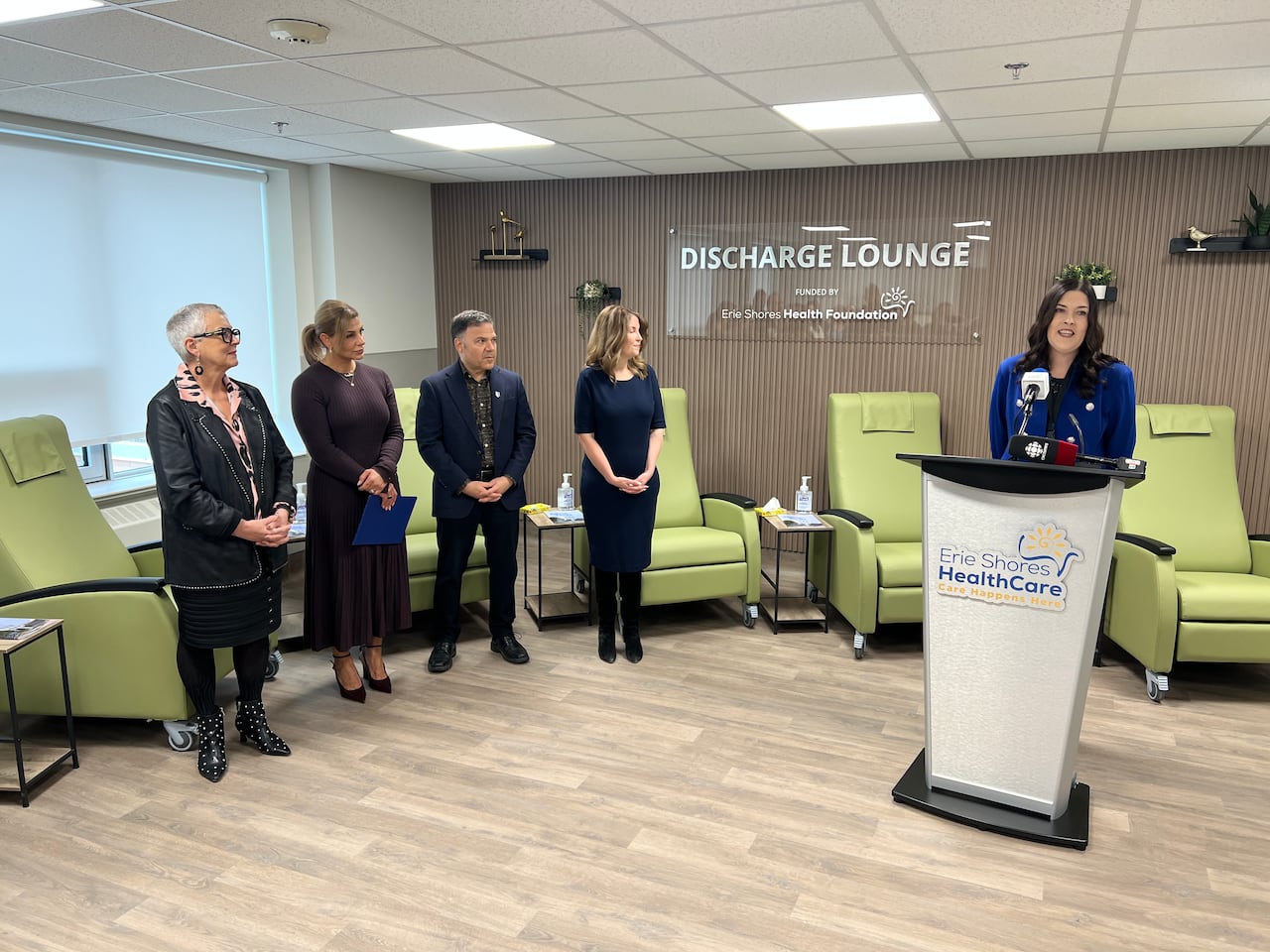 A woman stands at a podium with four other people standing to the side of her looking at her speak