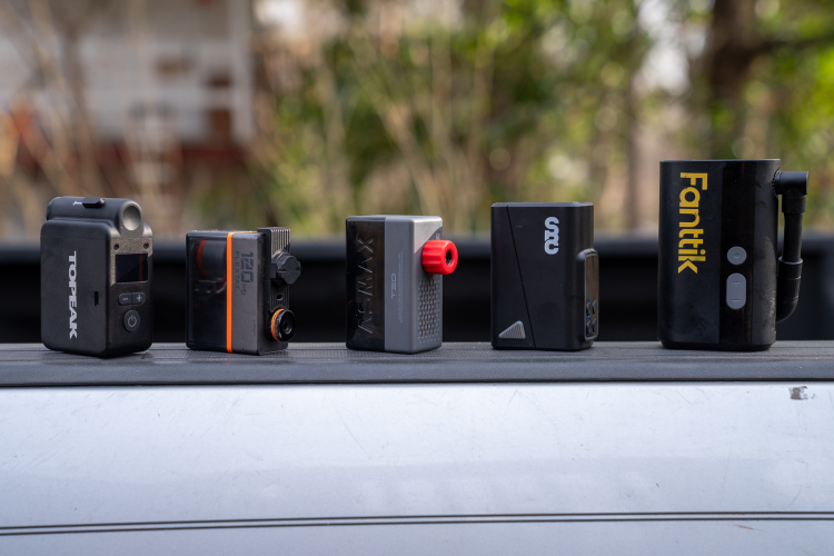 A lineup of five compact electronic devices placed on a flat surface. From left to right, they include a Topeak device, a black and orange Flexi unit, an XSIMAX product with a red knob, a small black device featuring a logo, and a larger Fanttik unit with a hose and buttons. The background shows blurred greenery, indicating an outdoor setting.
