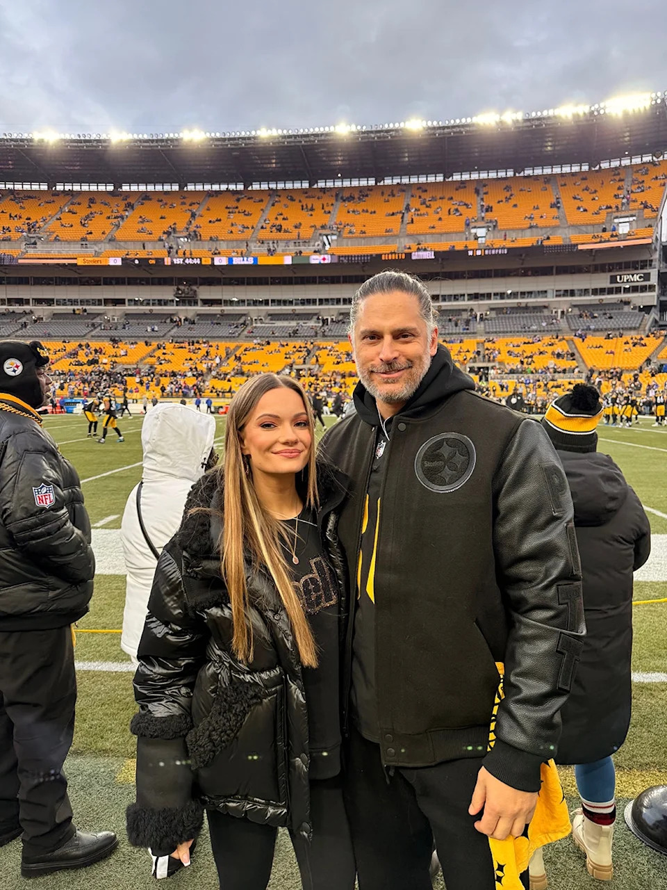 Joe Manganiello and Caitlin O'Connor smiling and wearing matching leather jackets standing on a football field.