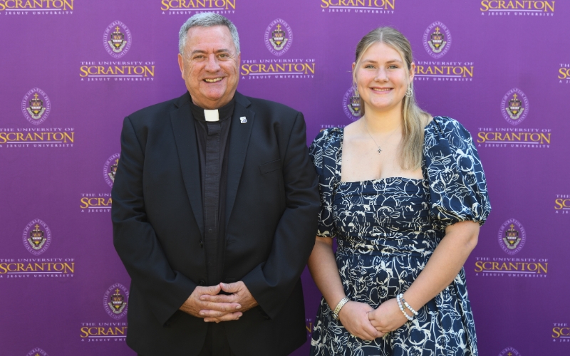 Two smiling individuals standing outdoors in front of a purple University of Scranton backdrop