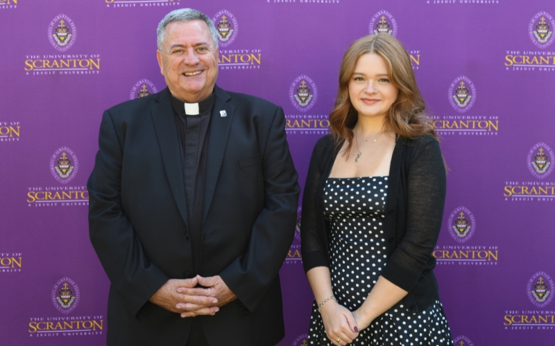 Two smiling individuals standing outdoors in front of a purple University of Scranton backdrop