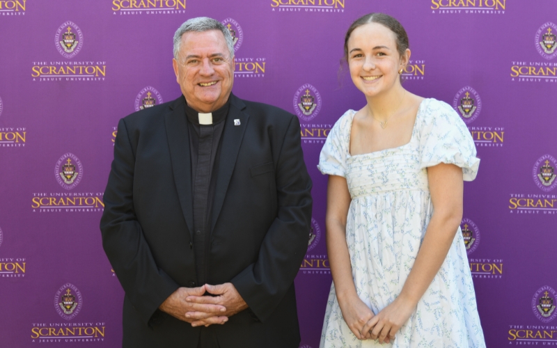 Two smiling individuals standing outdoors in front of a purple University of Scranton backdrop