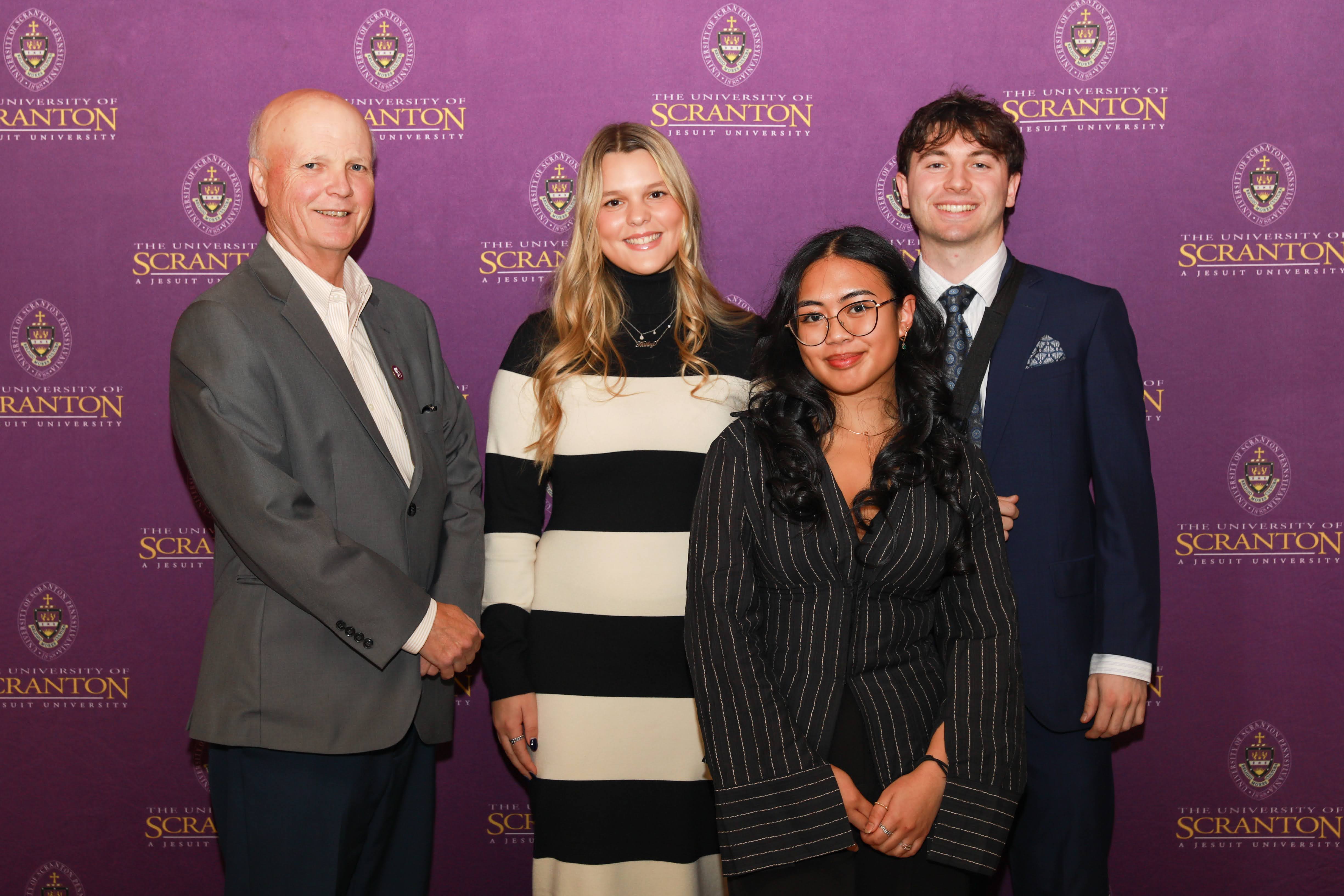 Four individuals pose for a photo together in front of a purple University of Scranton backdrop.