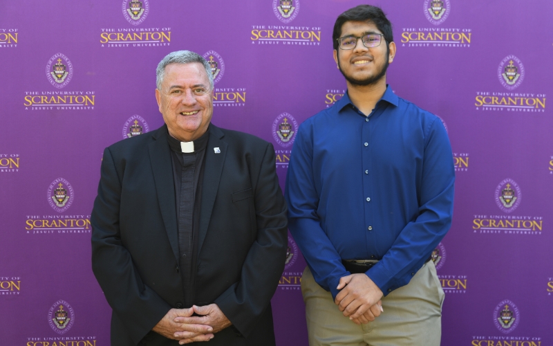Two smiling individuals standing outdoors in front of a purple University of Scranton backdrop