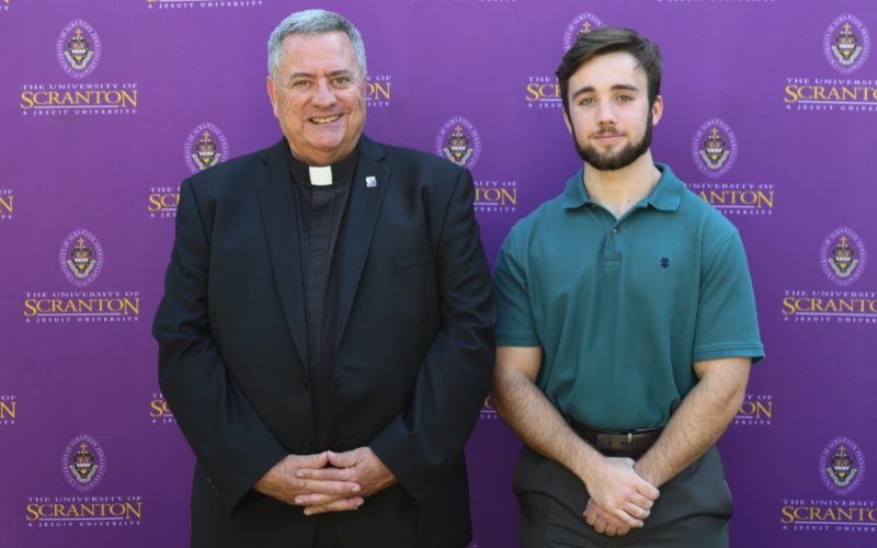 Two smiling individuals standing outdoors in front of a purple University of Scranton backdrop