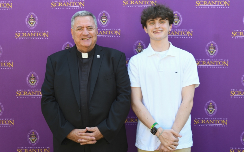 Two smiling individuals standing outdoors in front of a purple University of Scranton backdrop