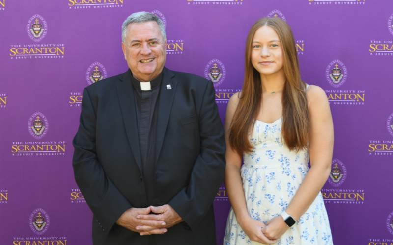 Two smiling individuals standing outdoors in front of a purple University of Scranton backdrop