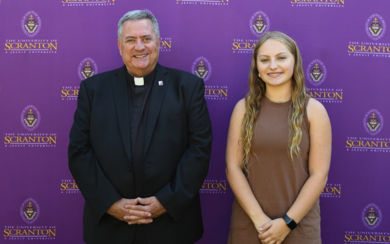 Two smiling individuals standing outdoors in front of a purple University of Scranton backdrop