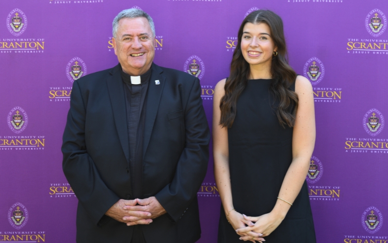Two smiling individuals standing outdoors in front of a purple University of Scranton backdrop