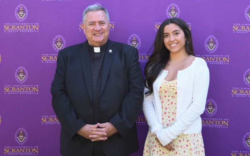 Two smiling individuals standing outdoors in front of a purple University of Scranton backdrop