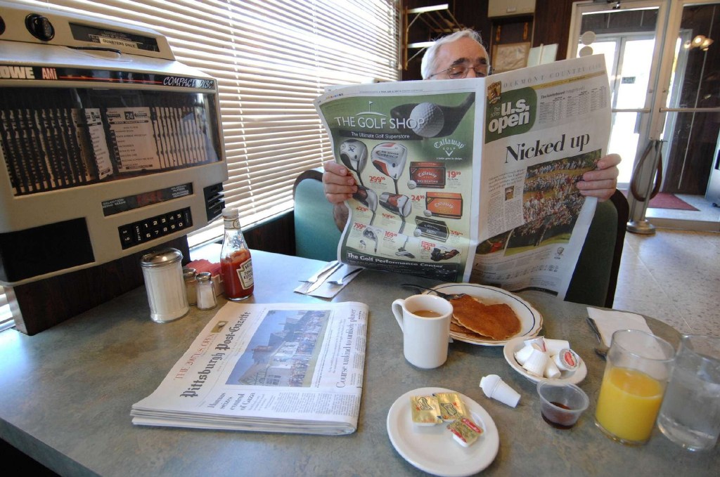 A man at a diner reads a newspaper with a "The U.S. Open" headline, with pancakes, coffee, and juice on the table.