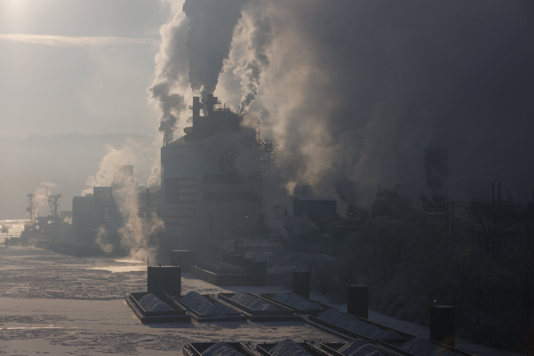 A large industrial facility emits thick smoke into the air, with smokestacks visible amid hazy, gray surroundings and a partially frozen body of water in the foreground.
