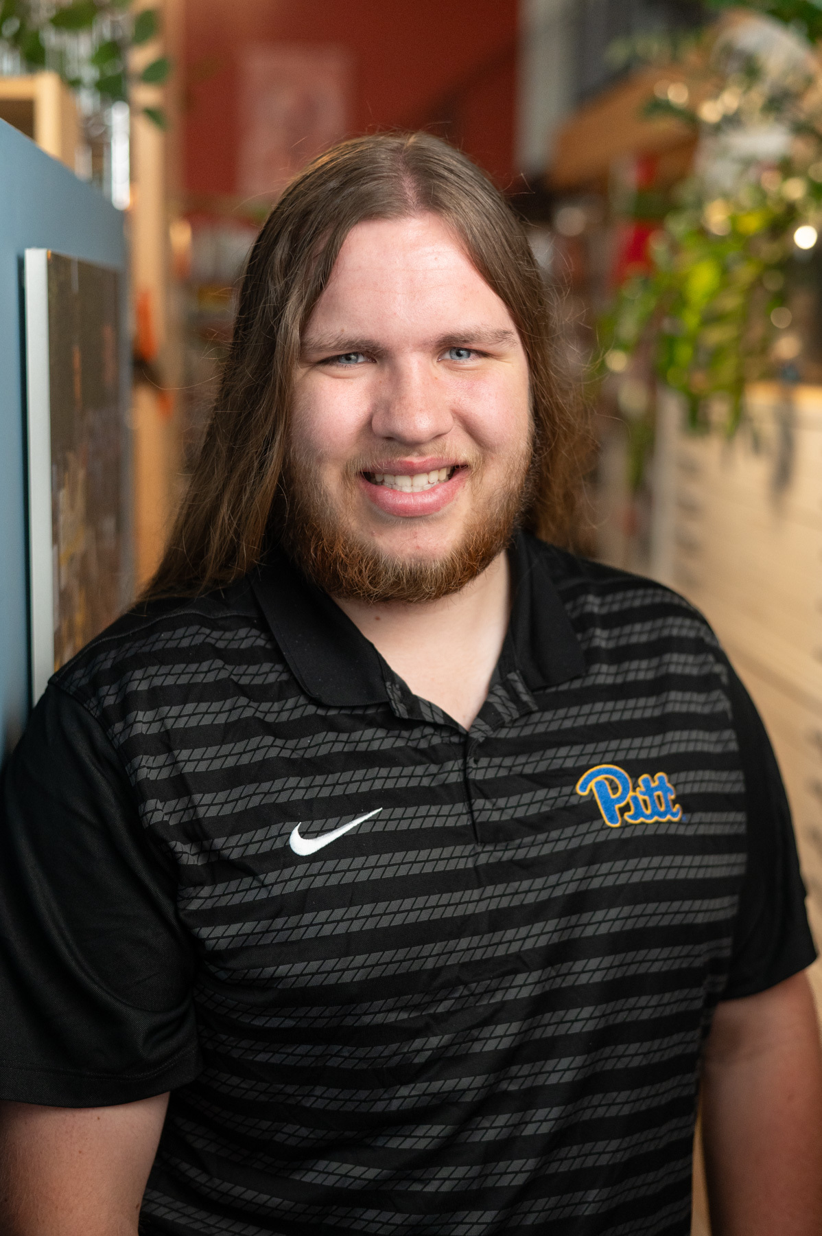 A person with long brown hair and a beard smiles at the camera, wearing a black Pitt Nike polo shirt, standing indoors next to shelves and plants.