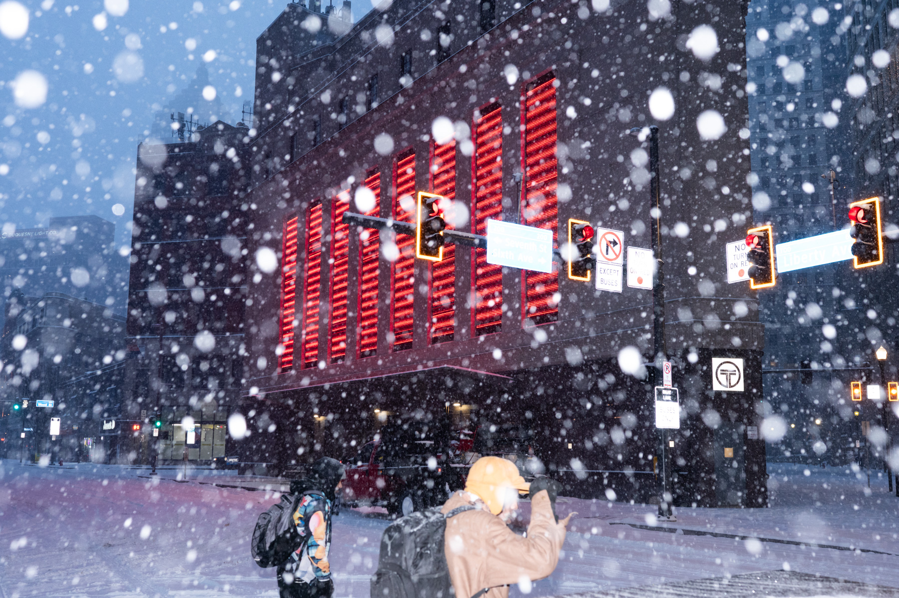 Two people walk in heavy snowfall near a city intersection with illuminated red lights on a building in the background. Traffic signals and snow-covered streets are visible.
