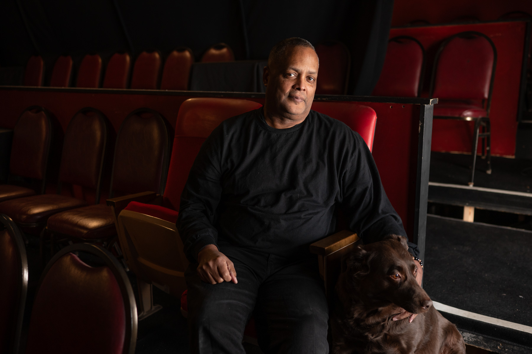 A man in a black shirt sits in a red theater seat with a brown dog by his side; empty chairs are visible in the background.