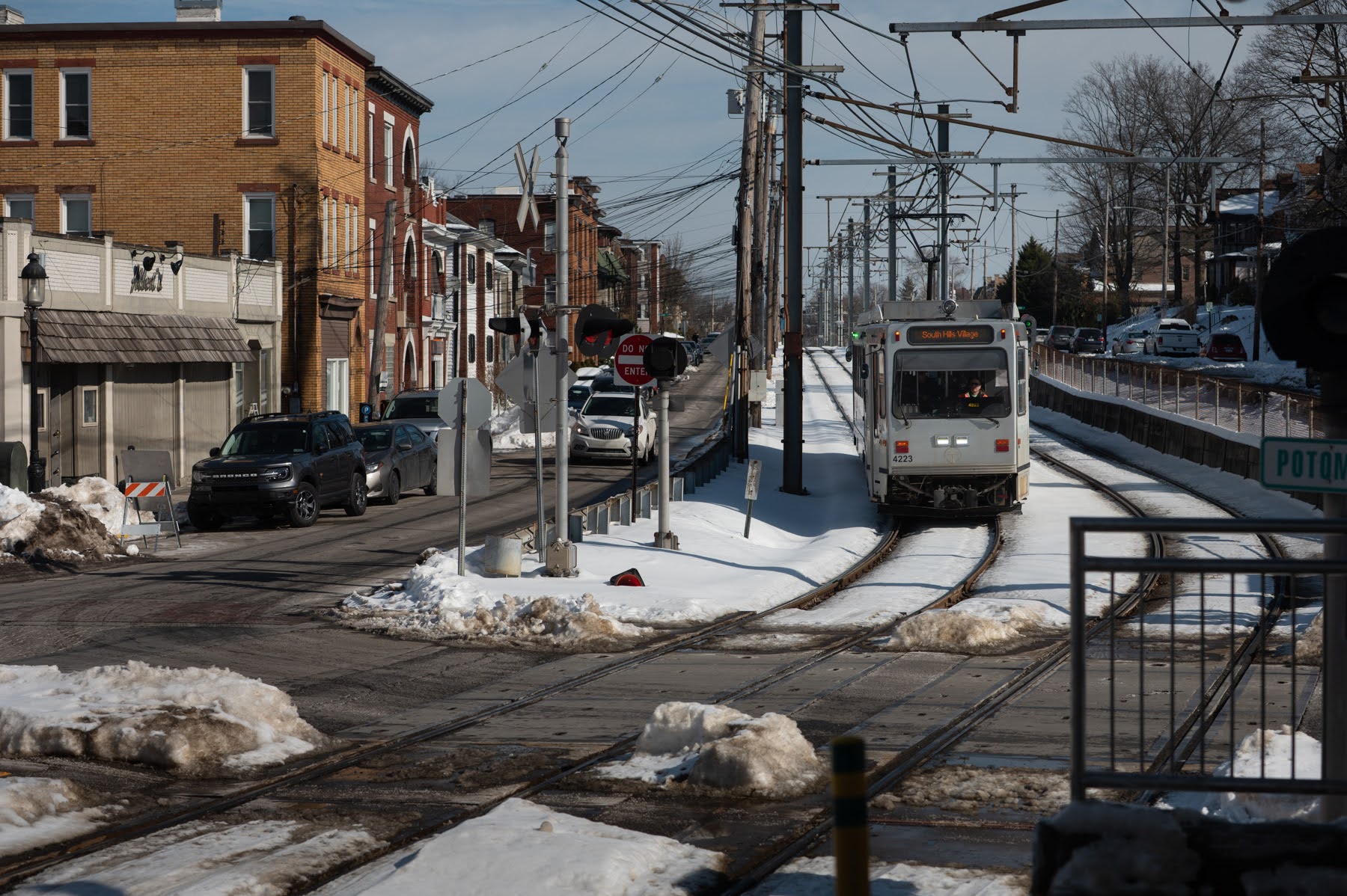 A light rail train approaches a level crossing in a snowy urban neighborhood, with cars waiting and brick buildings lining the street.