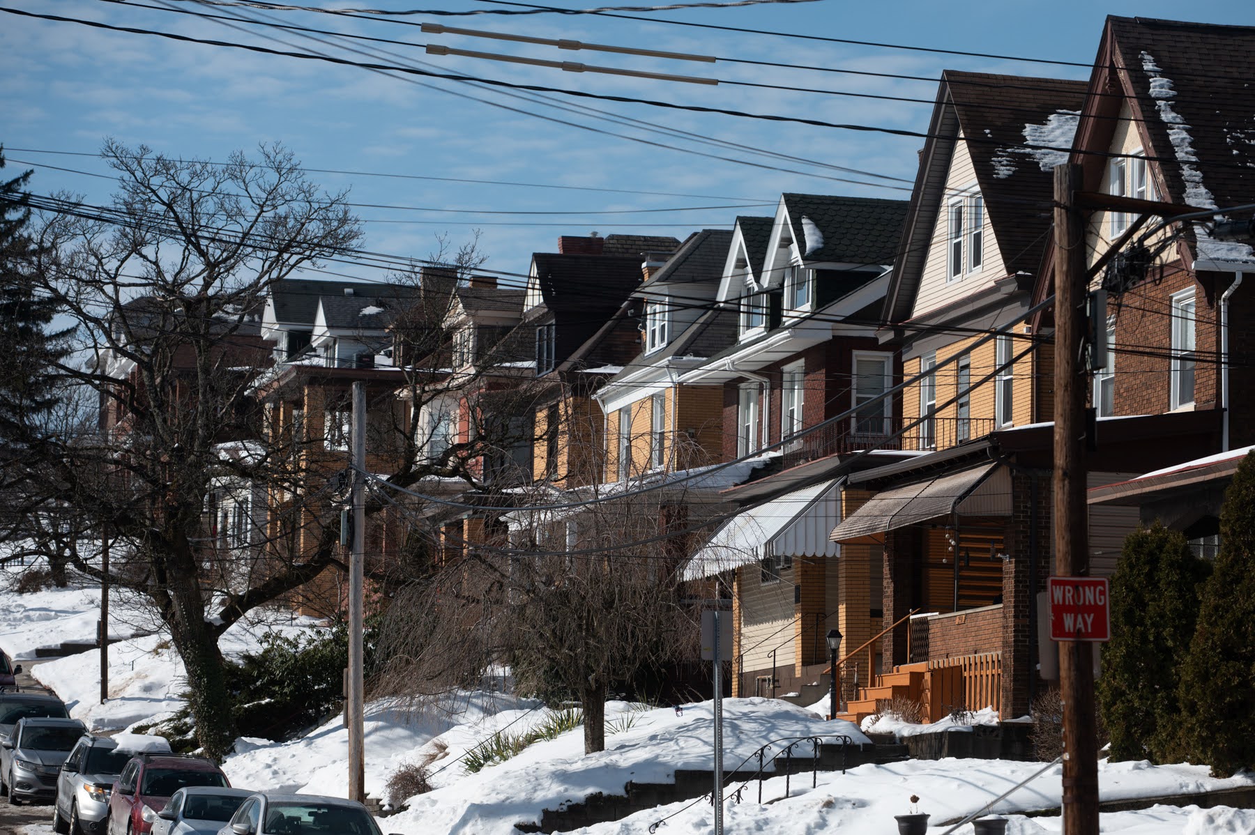 Row of houses on a snowy residential street with bare trees and power lines overhead, under a clear blue sky.