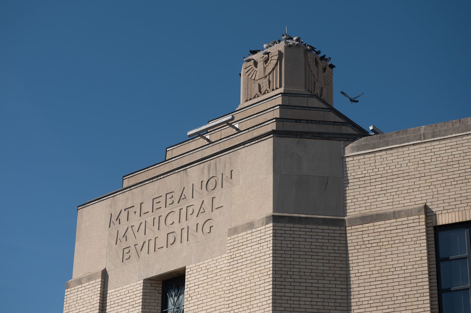 The upper section of the Mt. Lebanon Municipal Building with Art Deco architectural details, an eagle sculpture, and pigeons perched on the roof against a clear blue sky.