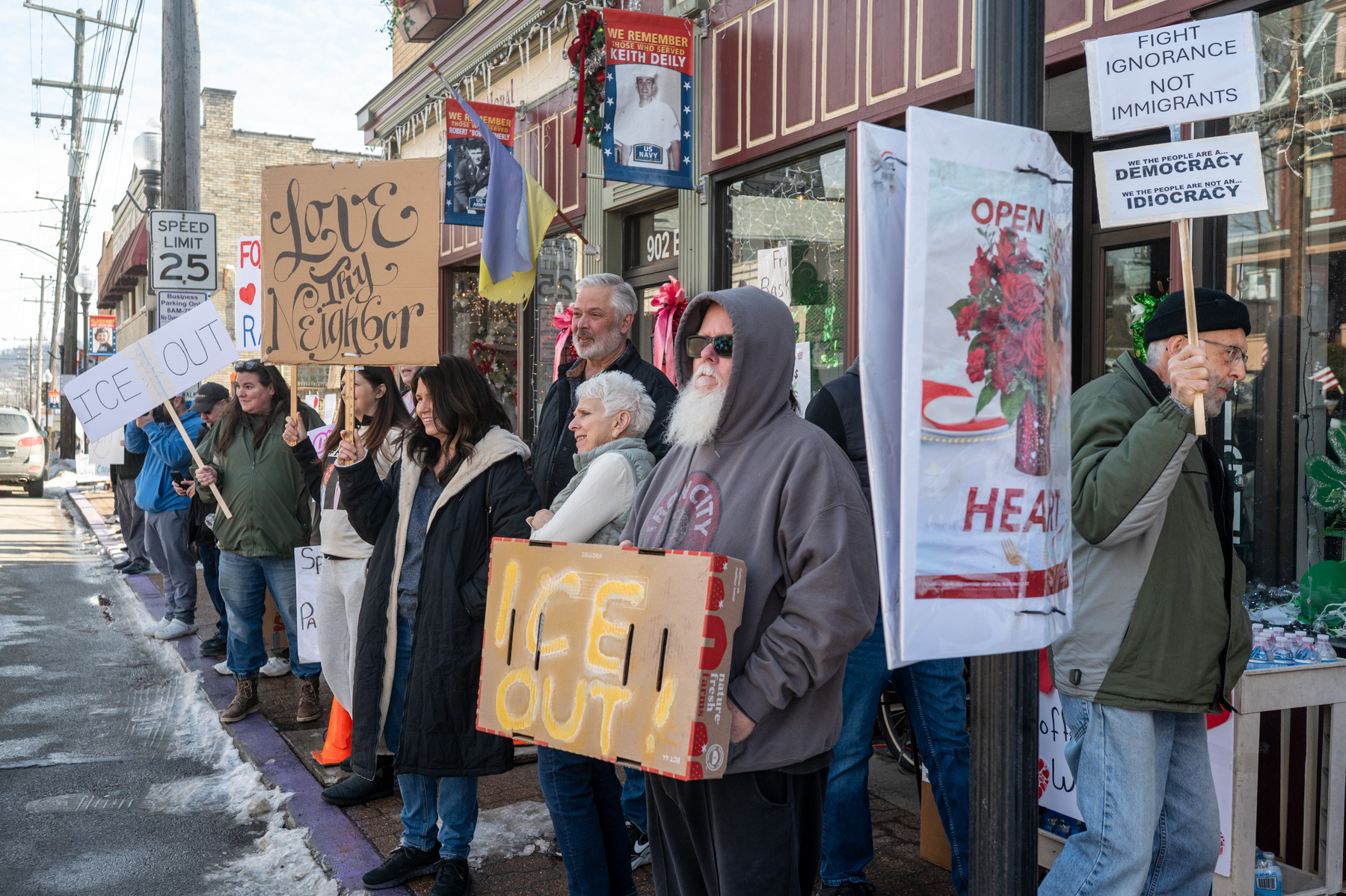 A group of people stand on a sidewalk holding protest signs with messages about immigration, democracy, and community support.