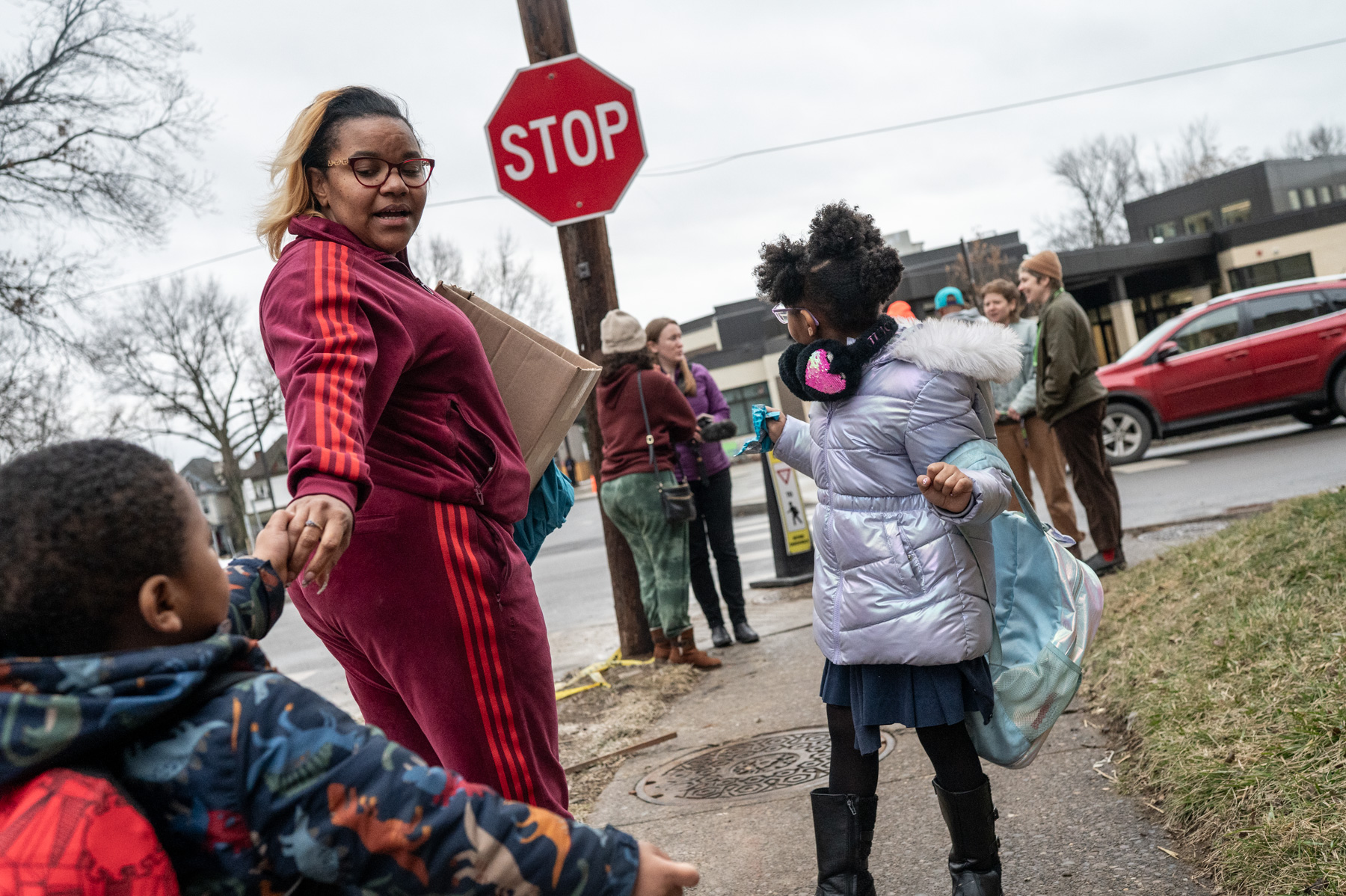 A woman in a red tracksuit holds a box and reaches for a child's hand near a stop sign; other people and a red car are in the background on a cloudy day.